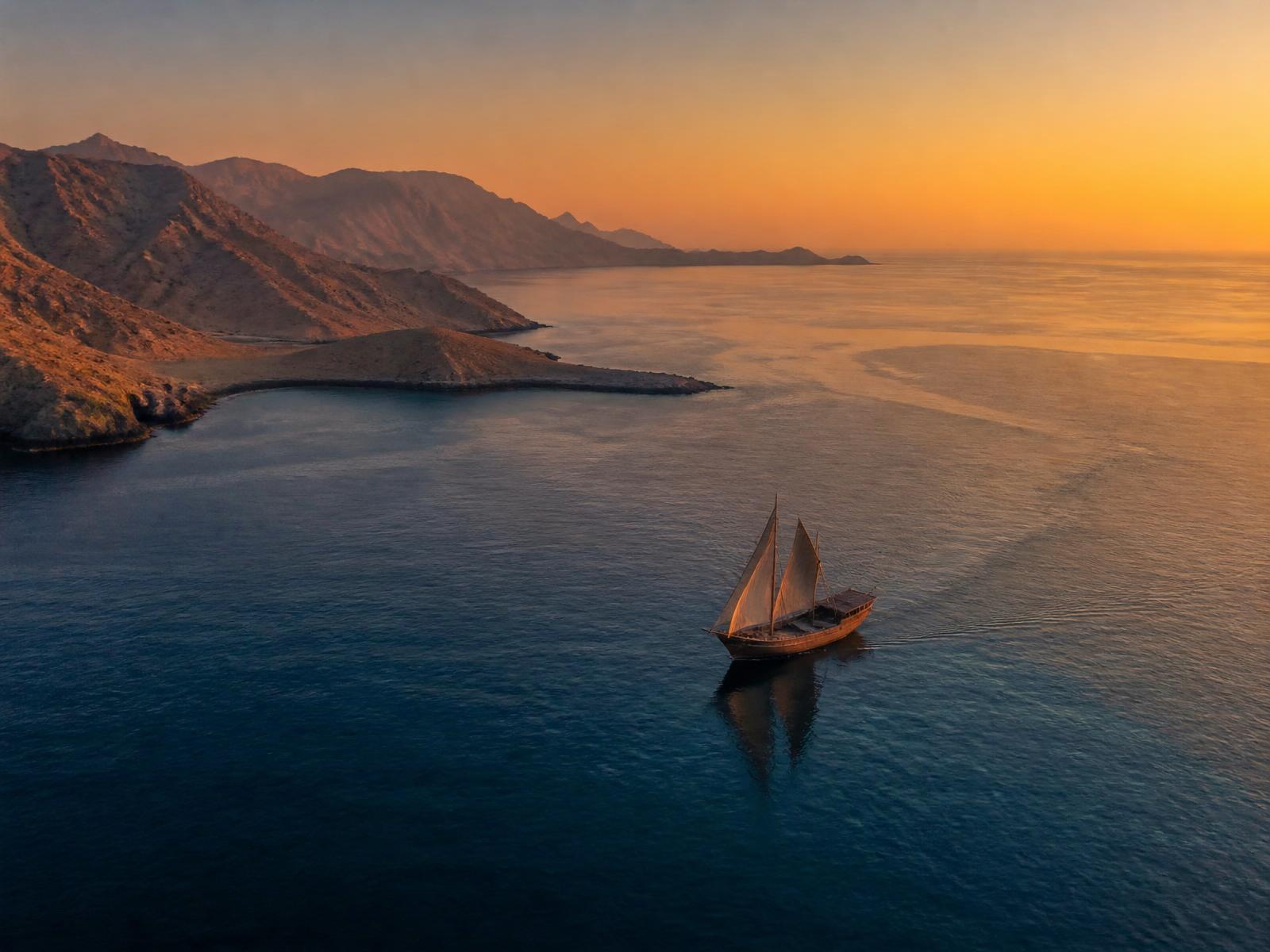 A single dhow sailing between Omani mountains and the open sea at sunset, symbolising the GCC–India corridor.